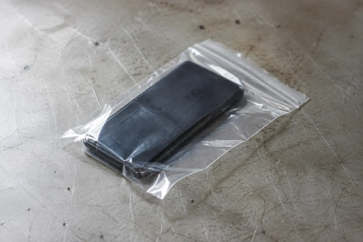 Černý telefon s vypnutým displejem leží v průhledném uzavíratelném sáčku na opotřebeném stole.